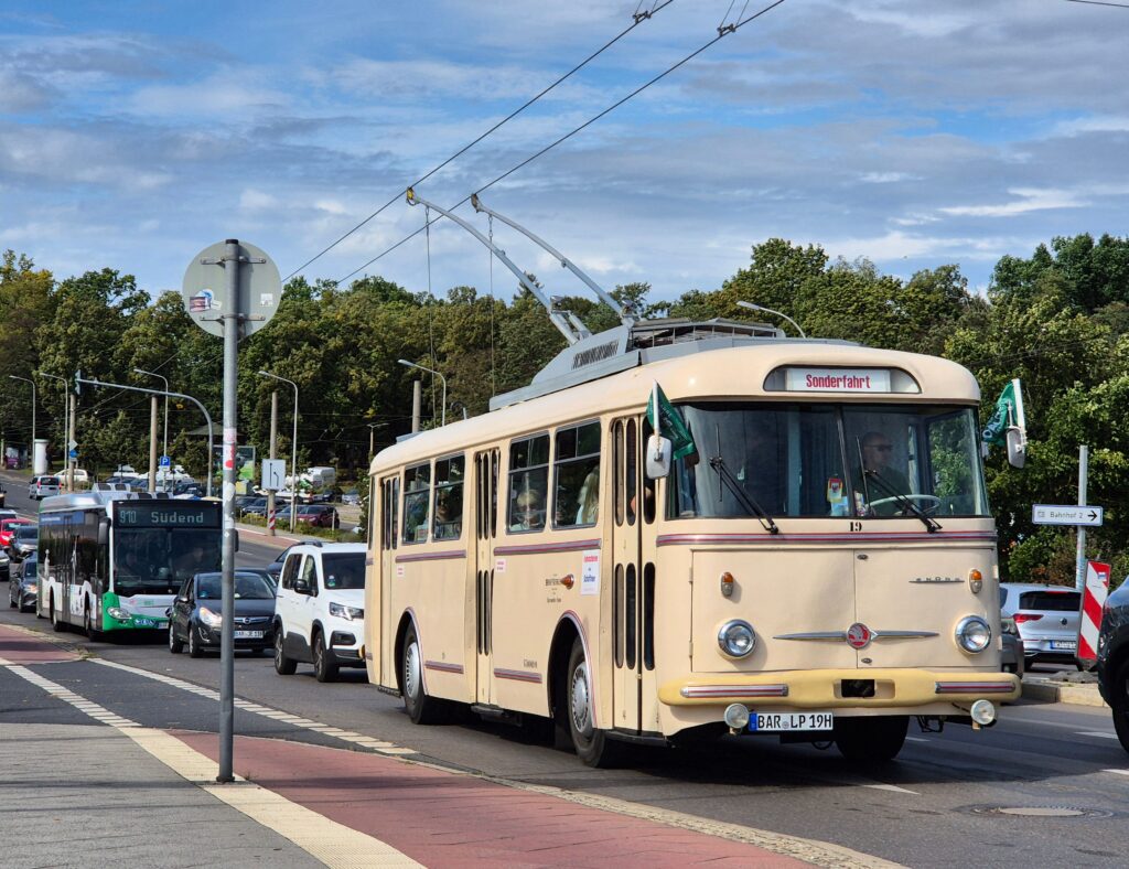 A classic trolleybus drives down a busy urban street lined with trees and modern vehicles.